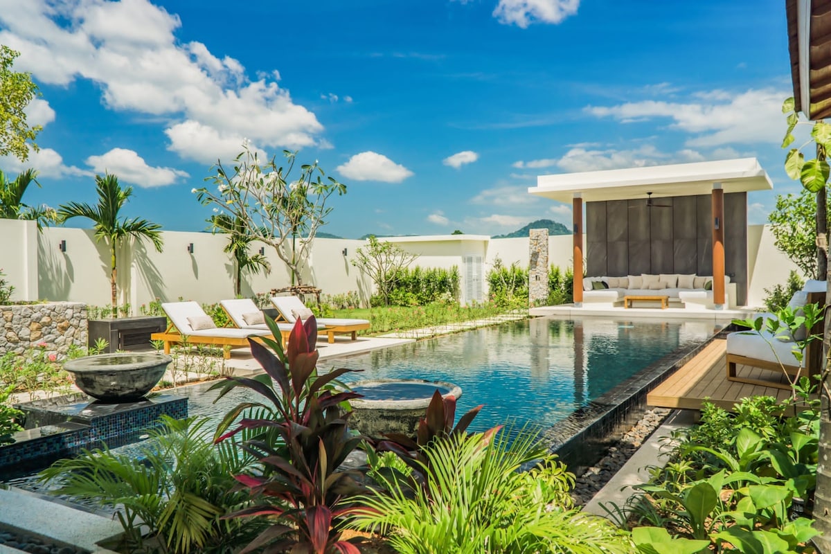 A serene outdoor setting features a large swimming pool surrounded by vibrant greenery and tropical plants. Sun loungers are positioned by the pool, while a covered seating area awaits nearby, providing space for relaxation and enjoyment of the tranquil landscape under a clear blue sky.