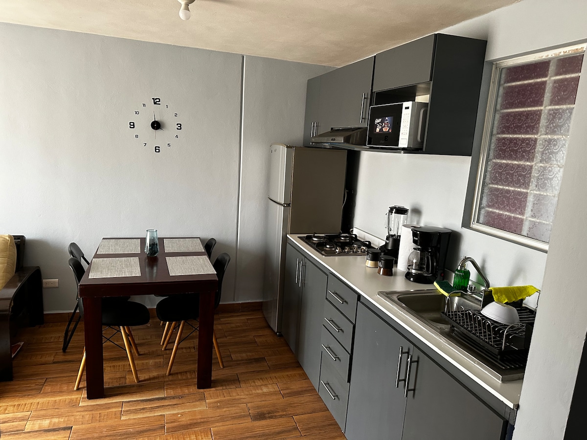 A modern kitchen space features sleek gray cabinetry and appliances, including a microwave and an oven. A dining table with four black chairs is positioned nearby, alongside a window allowing natural light. A sink and coffee maker are visible on the countertop, complemented by dishes in a drying rack.