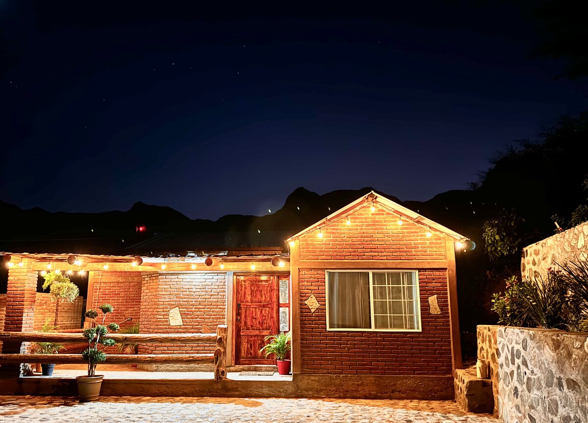 A brick house is depicted under a night sky, with warm string lights framing the entrance. A wooden door leads indoors, and a window is visible beside a small plant. The surrounding area includes stone pathways and distant mountains.