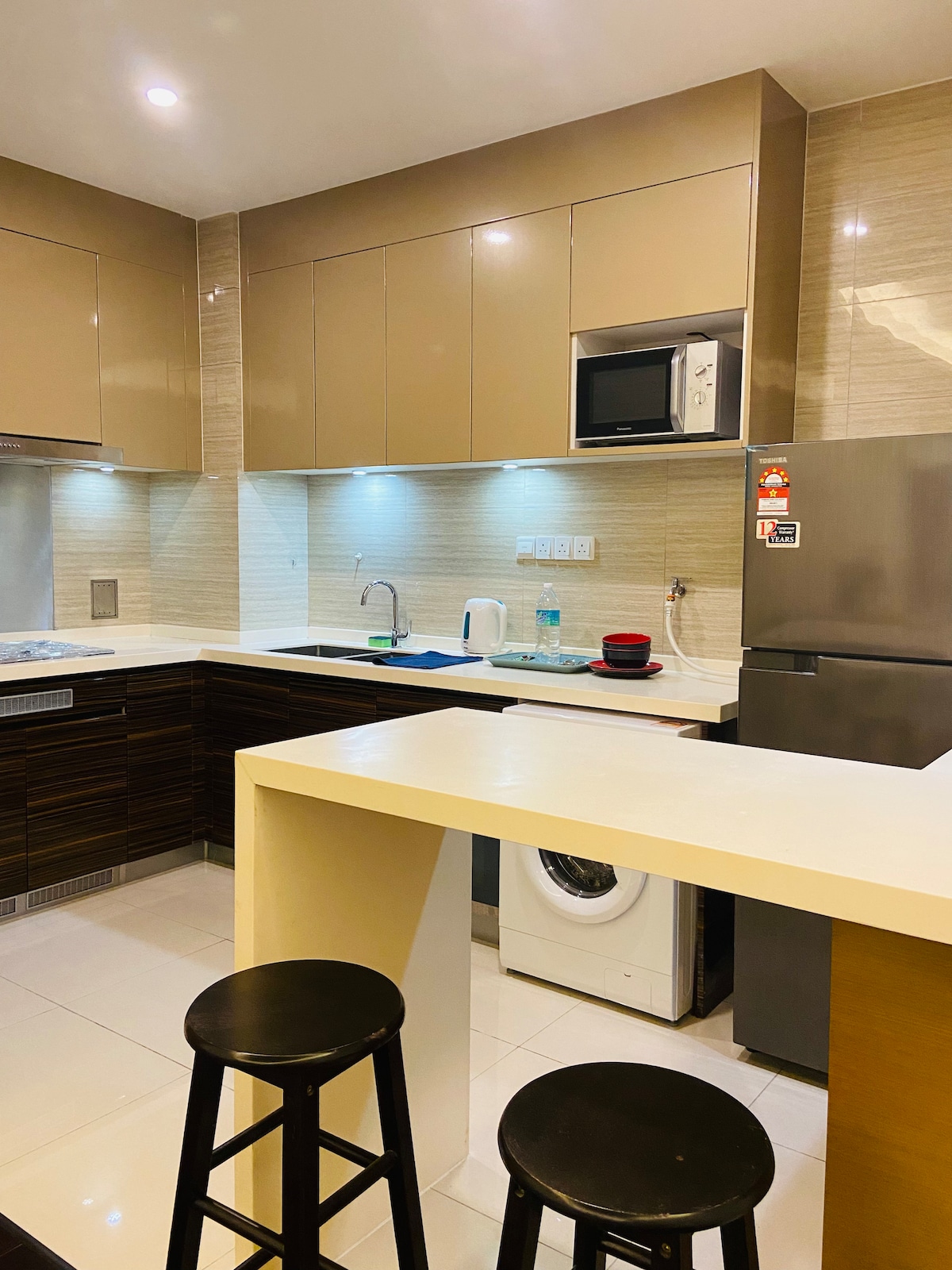 A modern kitchen area is equipped with sleek cabinetry and stainless-steel appliances. A compact breakfast bar with two stools provides seating. Under-cabinet lighting highlights the countertop, while a washing machine and refrigerator are positioned for convenience.