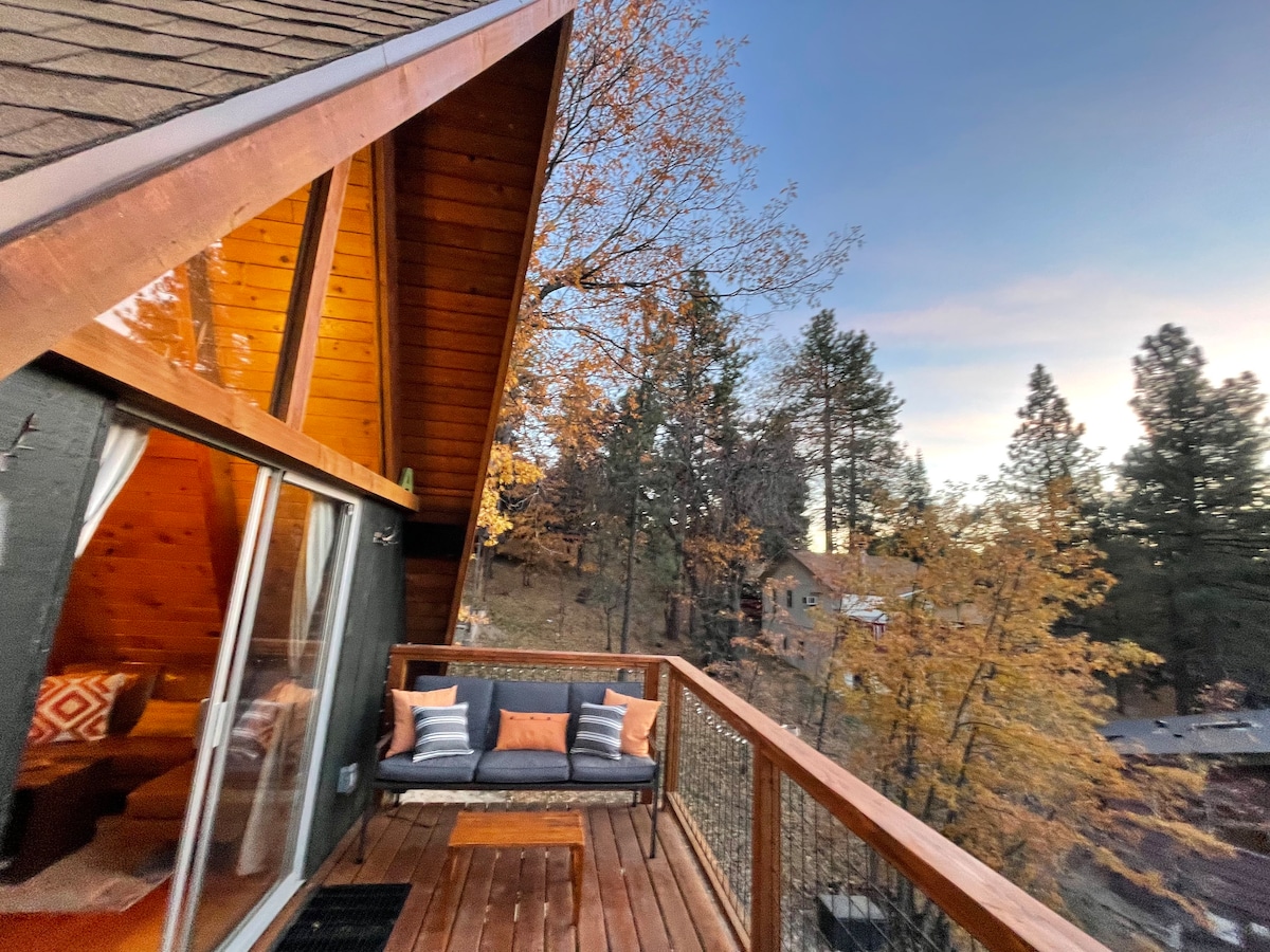 An outdoor deck features a seating area with cushions and pillows, surrounded by trees displaying autumn foliage. A large glass sliding door reflects the warm tones of the interior, creating a harmonious connection between indoor and outdoor spaces, while the A-frame structure rises above.
