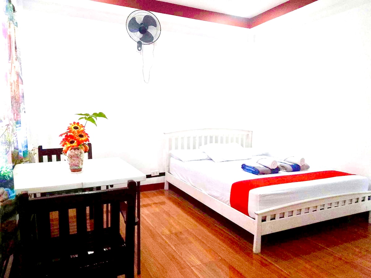 A bright sleeping area features a spacious bed with white linens and a vibrant red accent. A small dining table with two chairs is positioned next to the bed. Natural light brightens the room, complemented by a ceiling fan and a decorative vase of flowers.