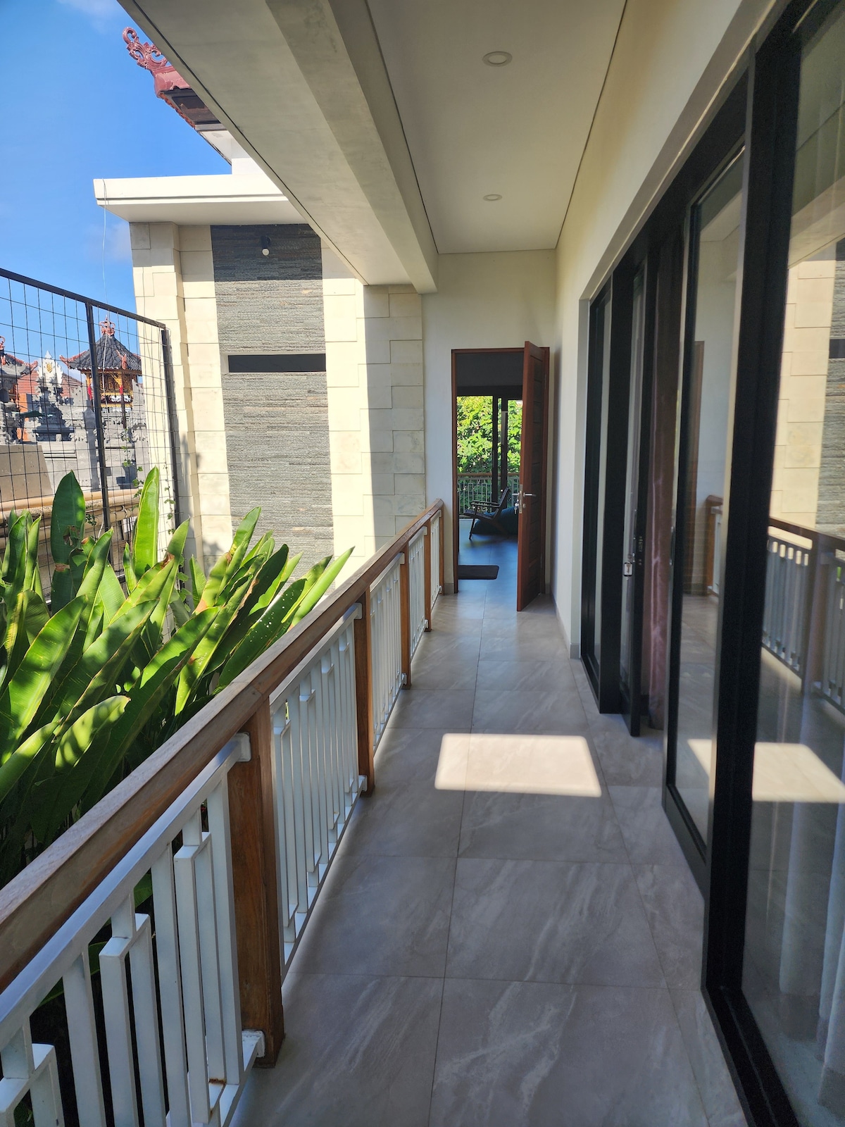 A bright hallway lined with large glass doors opens to a balcony overlooking greenery. The natural light enhances the spacious feel, while the smooth tiled flooring leads towards an inviting entryway.