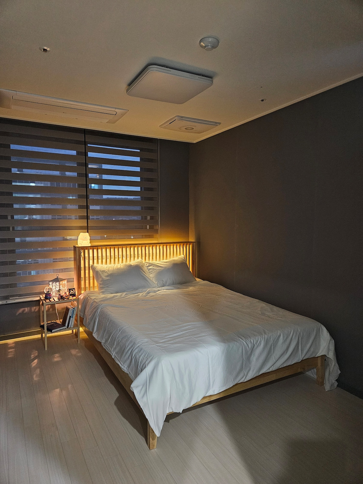 A queen-sized wooden bed is positioned against a dark wall, featuring crisp white bedding. Soft lighting emanates from a bedside table, which holds a small lamp and water bottles. Light filters through partially closed blinds, creating a calm ambiance in the room.
