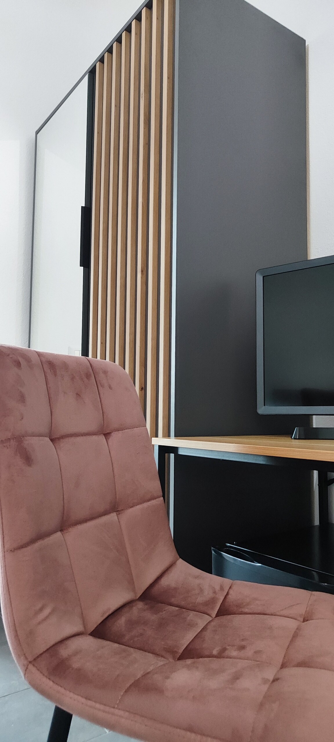 A modern workspace is featured, highlighting a sleek wooden desk and a plush chair with a soft pink hue. A flat-screen television is positioned next to the desk, while the backdrop includes stylish vertical wooden slats that add depth to the design.