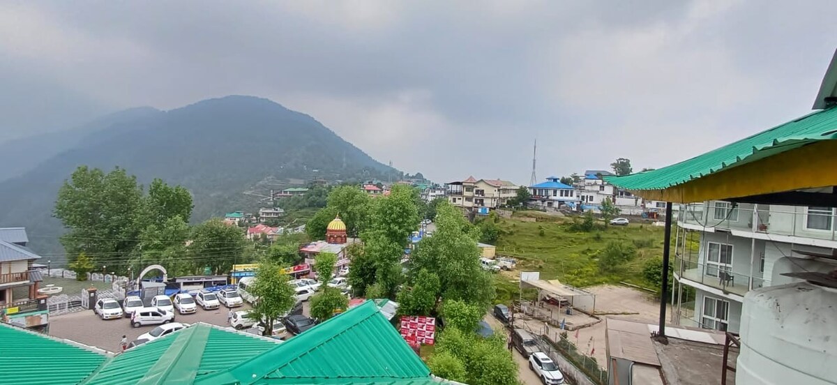 A panoramic view captures the surrounding hills and the quaint village. Various buildings are visible, along with green spaces and parked vehicles. The sky is overcast, enhancing the serene landscape of the area, typical of the Dharamshala region.