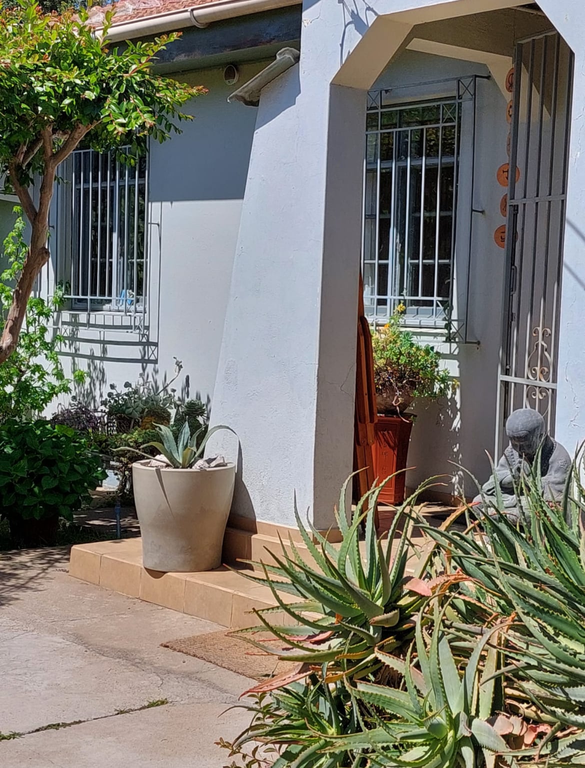 A serene entrance area is framed by lush greenery, featuring a prominent potted plant and a decorative statue. The walkway is lined with a blend of natural stone and concrete, leading to a white wall with barred windows, enhancing the tranquil ambiance.