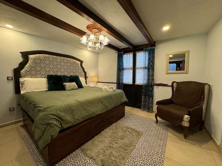 Amplia recámara con cama king size, techo de vigas de madera y lámpara de cristal, que combinan el estilo colonial con un diseño elegante y contemporáneo. Incluye baño privado y detalles de autor que crean un ambiente acogedor y sofisticado.
