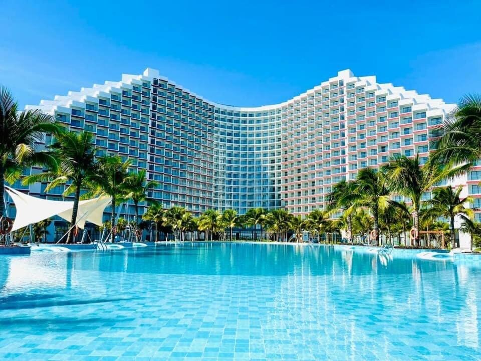 A modern resort building features a wave-like design, surrounded by lush palm trees. A large shimmering pool stretches in the foreground, reflecting the clear blue sky and enhancing the serene environment.