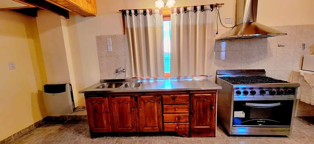 A functional kitchen space features wooden cabinetry and a stainless steel stove with a hood. A double sink is positioned beneath a window draped with light-colored curtains. The floor is covered in gray tiles, contributing to the serene atmosphere.