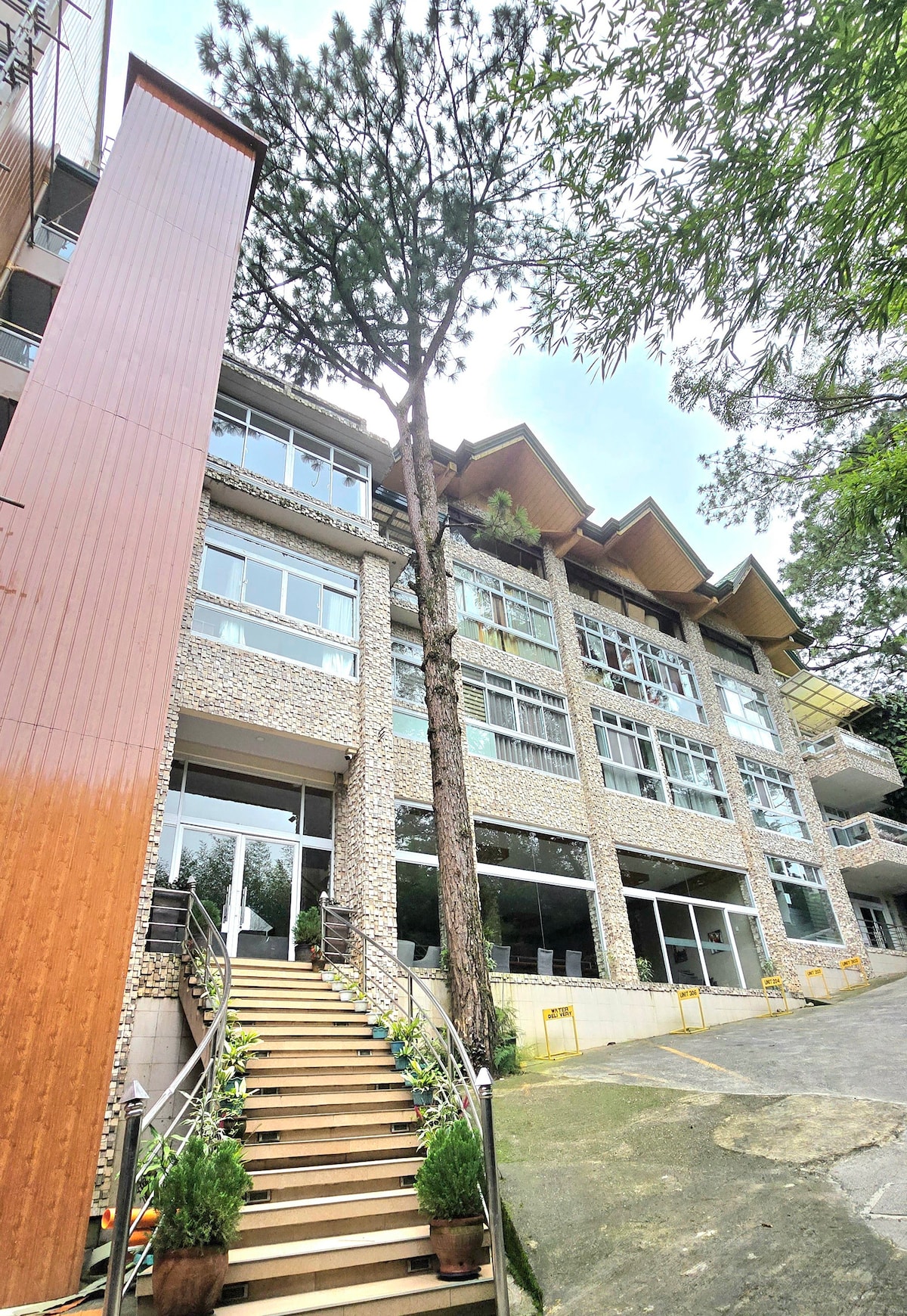 The exterior of the condo displays a modern three-story structure featuring large glass windows and a distinctive wooden entryway. A tall tree is positioned adjacent to the building, contributing to the surrounding greenery, while landscaped steps lead to the main entrance.