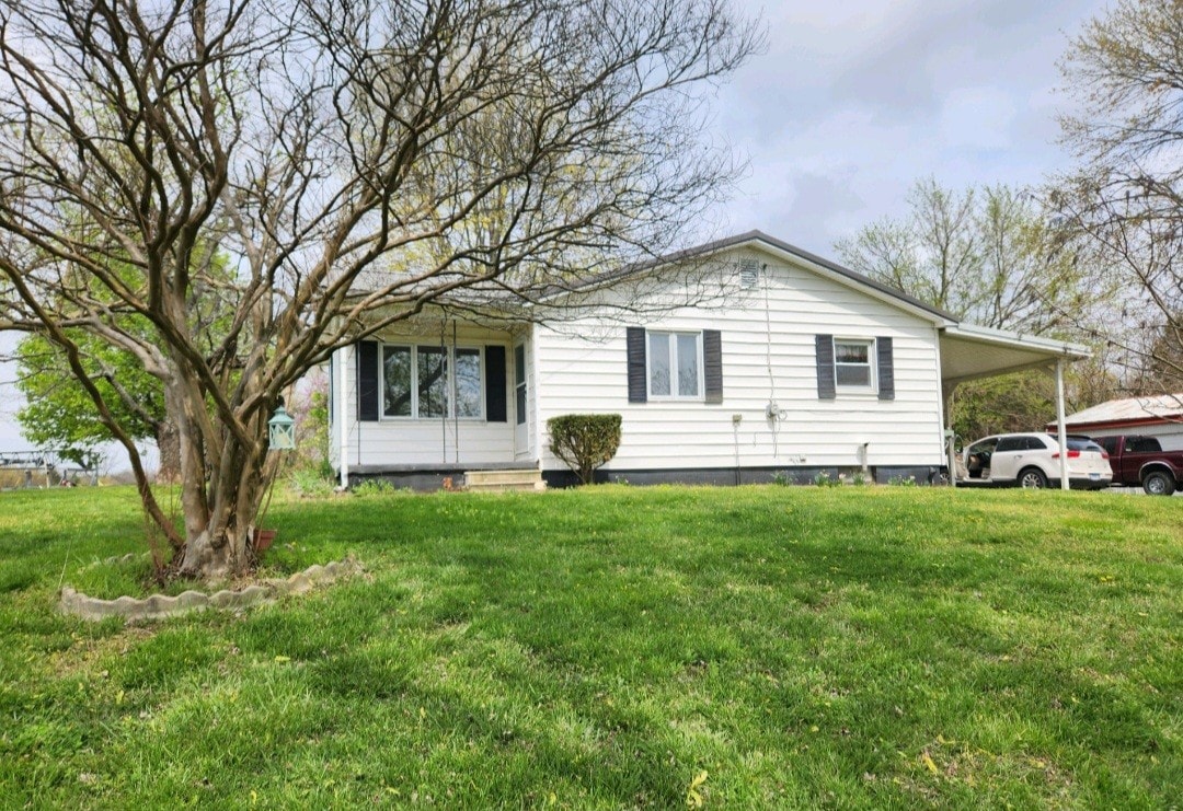 A single-story farmhouse is set against a backdrop of greenery, showcasing white siding and blue shutters. A covered porch extends along the front, while a tree provides shade in the yard. The surrounding area features well-maintained grass, enhancing the inviting exterior.