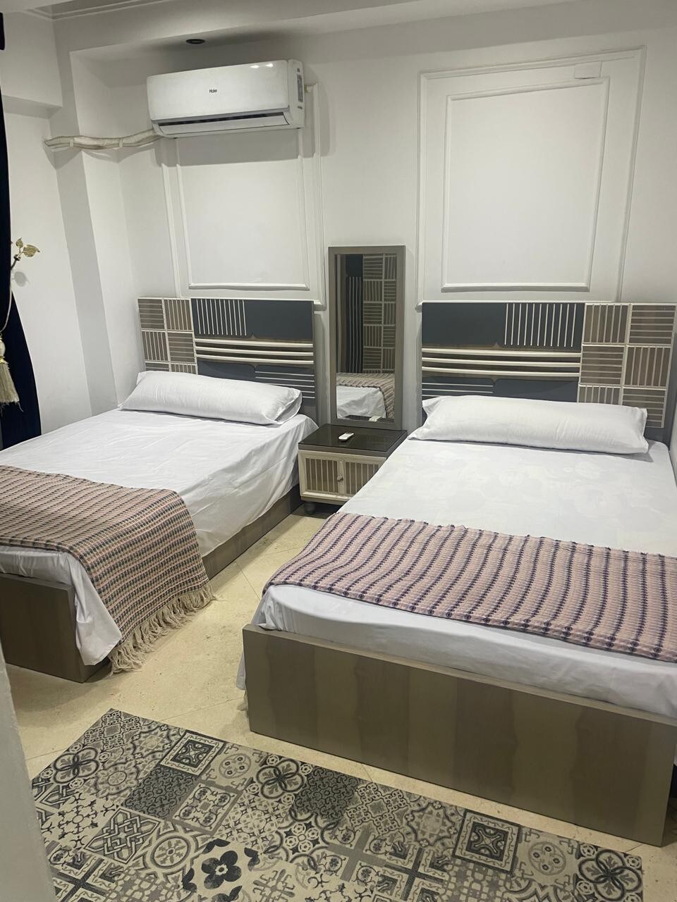The image showcases a room with two spacious beds, each adorned with light bedding and a decorative throw. A mirrored wardrobe is situated between the beds, reflecting the light in the airy space. Decorative tiles cover the floor, adding a subtle pattern.