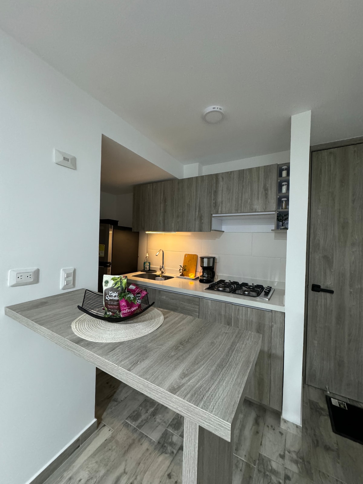 A modern kitchen area features light wood cabinetry and a minimalist design. An efficient stovetop and a coffee maker are visible on the countertop. Soft lighting creates a welcoming ambiance, complemented by decorative elements on a woven placemat.