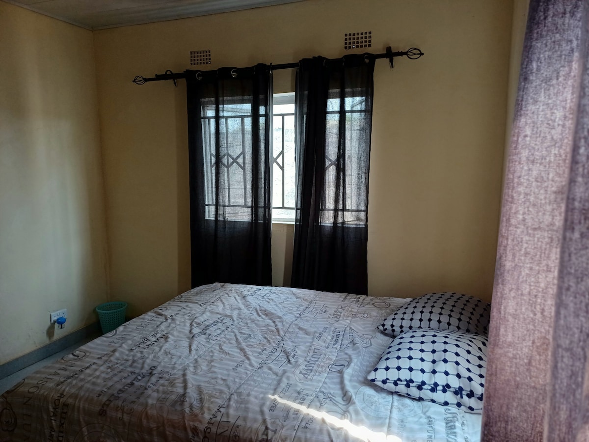 A cozy sleeping area is presented with a spacious bed covered in light bedding. Two patterned pillows are arranged neatly on the bed. Natural light filters through a window, partially obscured by dark curtains, illuminating the warm, neutral walls.