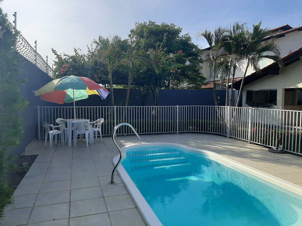 A private swimming pool is framed by a patio adorned with colorful umbrellas and white chairs. Lush greenery and flowering plants enhance the outdoor ambiance, while a secure fence ensures privacy. The inviting pool area is bathed in natural light, providing a perfect space for relaxation.