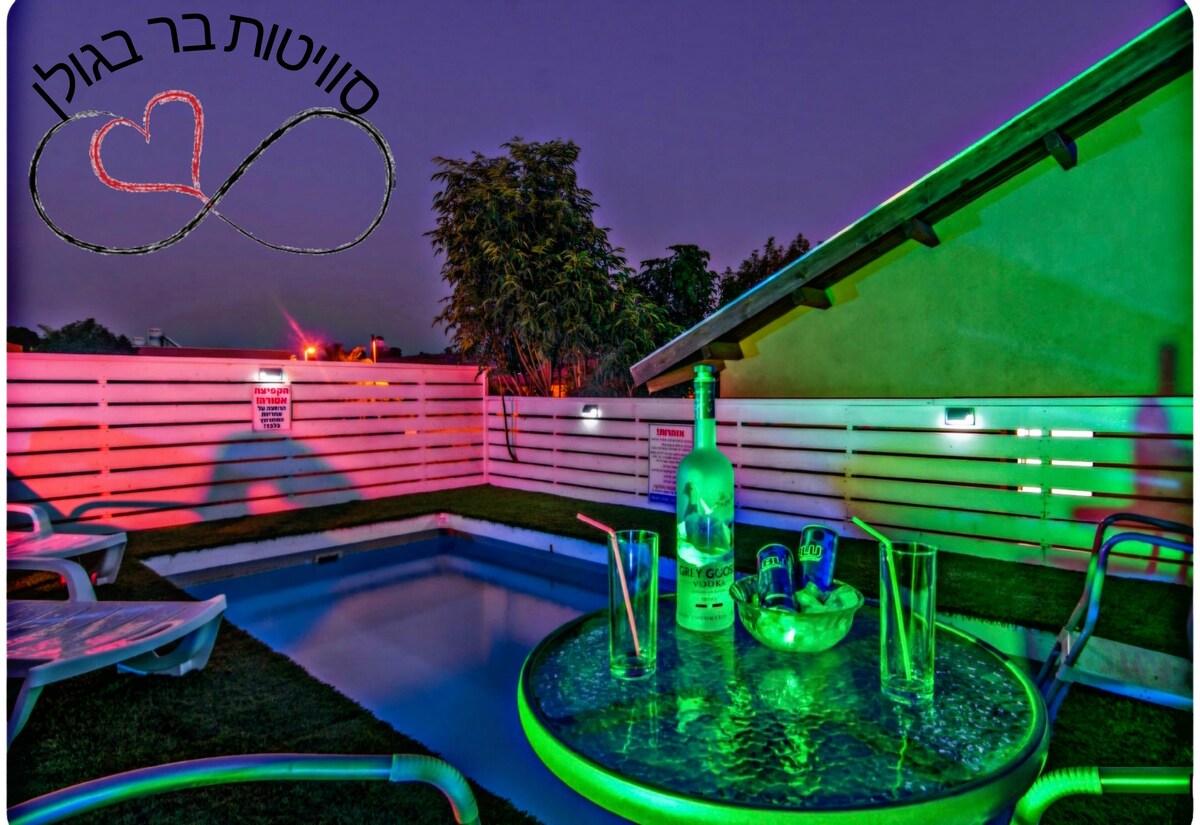 A small outdoor area is presented, featuring a refreshing pool surrounded by green grass. Colorful lighting enhances the space, and a table is set with refreshing drinks and glasses. Lounge chairs are positioned nearby, creating a relaxing setting under a twilight sky.
