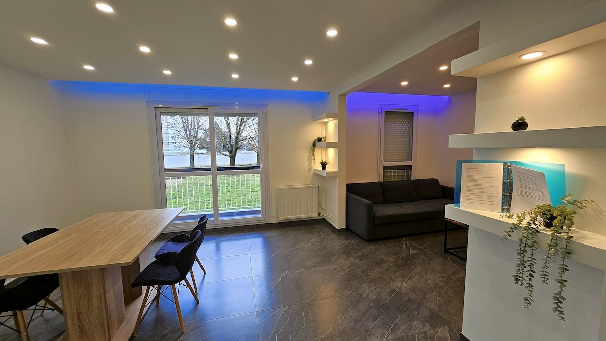 A modern living area showcases a spacious dining table surrounded by black chairs. Soft blue accent lighting complements the natural light coming through large windows, which offer a view of greenery outside. A comfortable sofa and stylish shelving enhance the contemporary decor.