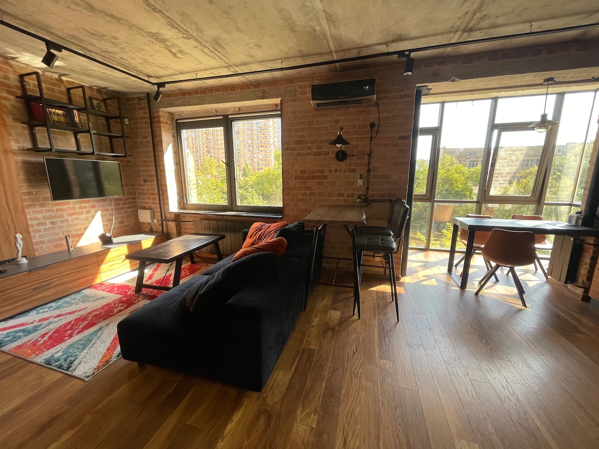 The living area showcases natural light streaming through large panoramic windows, illuminating the wooden floor. A comfortable dark sofa is paired with a vibrant rug. A sleek dining table with chairs enhances the space, complemented by a wall-mounted television.