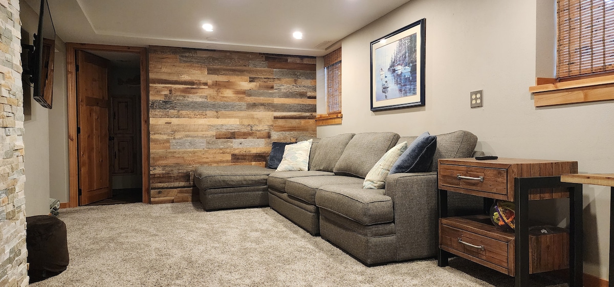 A cozy living area is presented with a gray sectional sofa and a wooden side table. Warm light illuminates the space, highlighting the textured wooden accent wall. A blend of natural tones is complemented by a soft carpet underfoot, enhancing comfort.