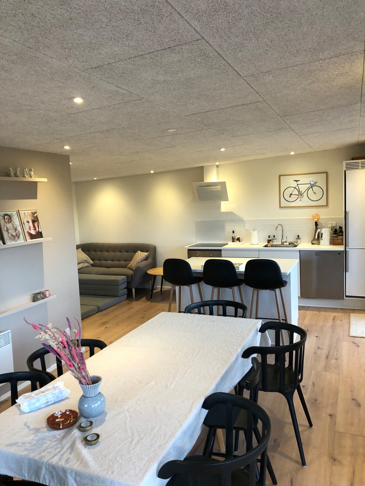 A modern living area features a comfortable gray sofa and a sleek kitchen counter with bar stools. A dining table is set with simple decor, surrounded by black chairs, while shelves display tasteful items. Soft lighting enhances the airy and inviting environment.