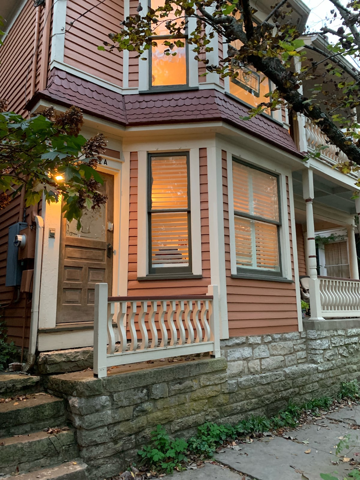 An exterior view of a Victorian home features warm lighting glowing through the windows. The entrance showcases a wooden door adjacent to a staircase leading to the front porch, which is bordered by a decorative railing and complemented by stone steps.