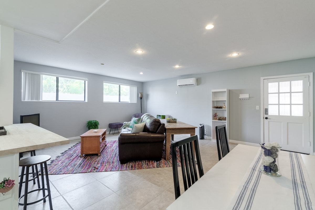 A spacious living area is presented with ample natural light from multiple windows. A comfortable sofa faces a coffee table, alongside a decorative area rug. A dining table with high-backed chairs is visible in the foreground, while a door leads to the exterior.