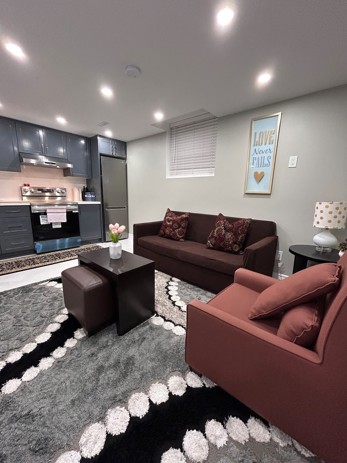 A cozy living area features two comfortable seating options, each with decorative pillows. A small coffee table is positioned between them. In the background, the kitchen area is visible, showcasing modern cabinetry and appliances. Soft lighting accentuates the warm tones of the furnishings.