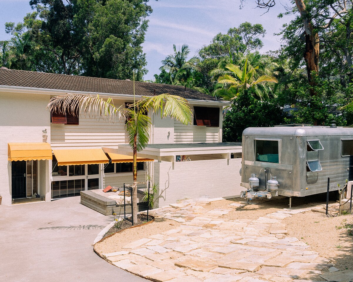 The exterior showcases a spacious family home featuring cream-colored siding and large windows. A vintage silver trailer is parked nearby, complemented by palm trees and vibrant yellow awnings. A stone pathway leads to a deck area, surrounded by greenery and a peaceful atmosphere.