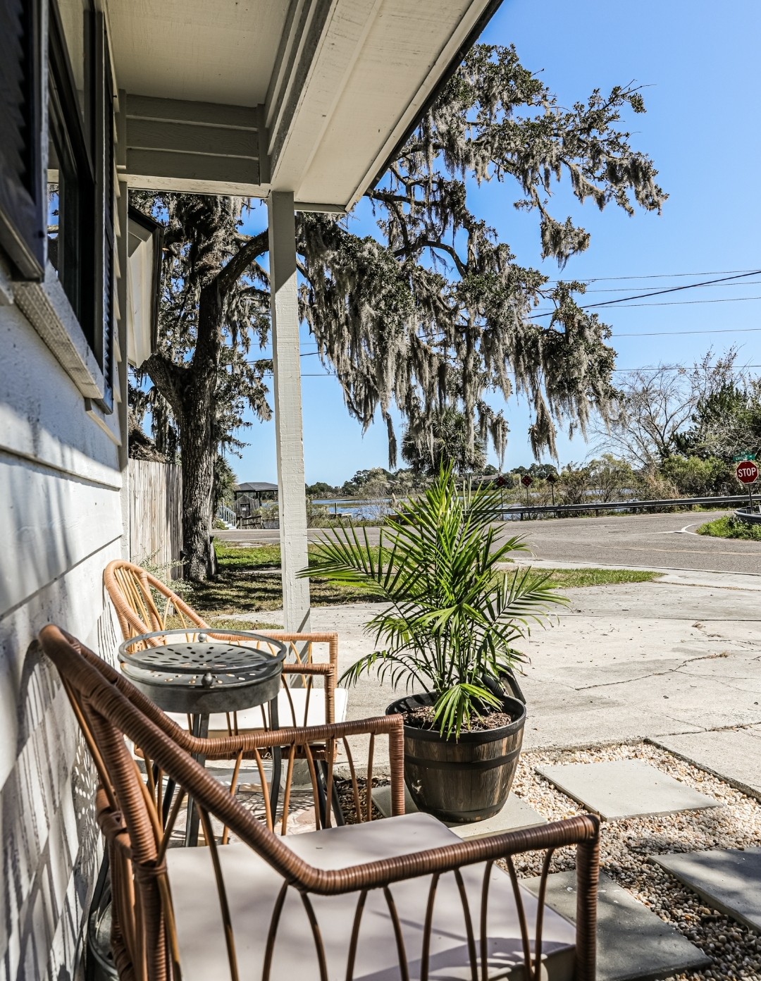 A charming outdoor seating area is featured, with two wicker chairs and a small table positioned beside a potted palm. Spanish moss drapes from nearby trees, and a view of the road is visible in the background under clear blue skies.