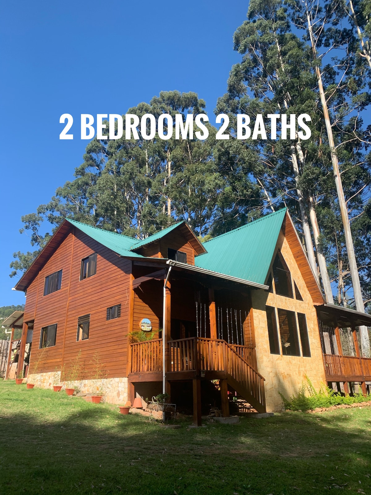 A two-story cabin, primarily constructed of wood, is set against a backdrop of tall trees. The structure features a green metal roof and a spacious front porch, providing a welcoming entrance to the home.