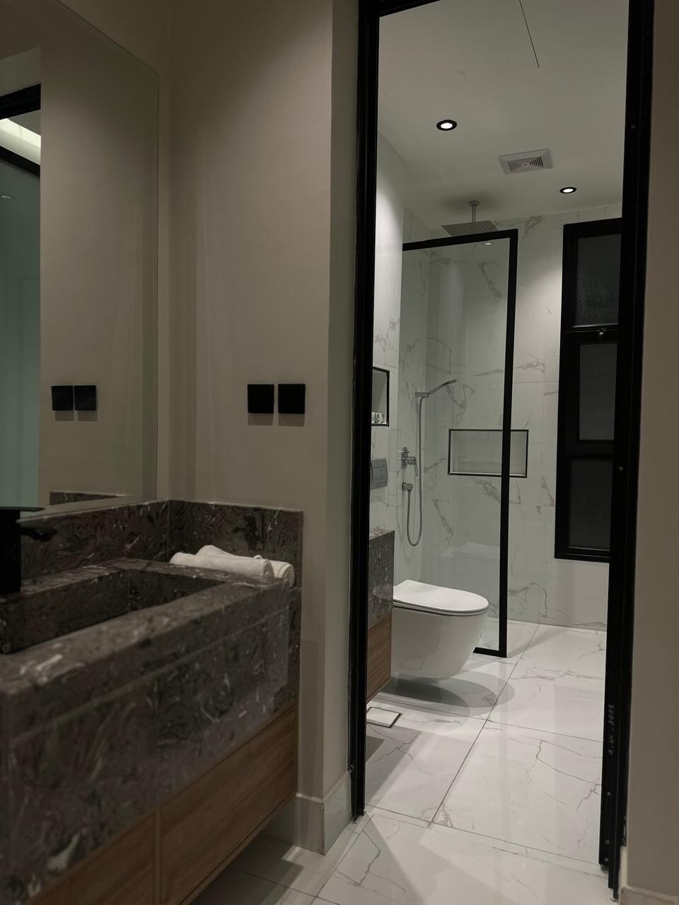 A modern bathroom is featured, showcasing a sleek, stone countertop with a built-in sink. A walk-in shower with glass doors and contemporary fixtures is visible. The space is illuminated by recessed lighting, enhancing the neutral color palette of the large tiles and walls.
