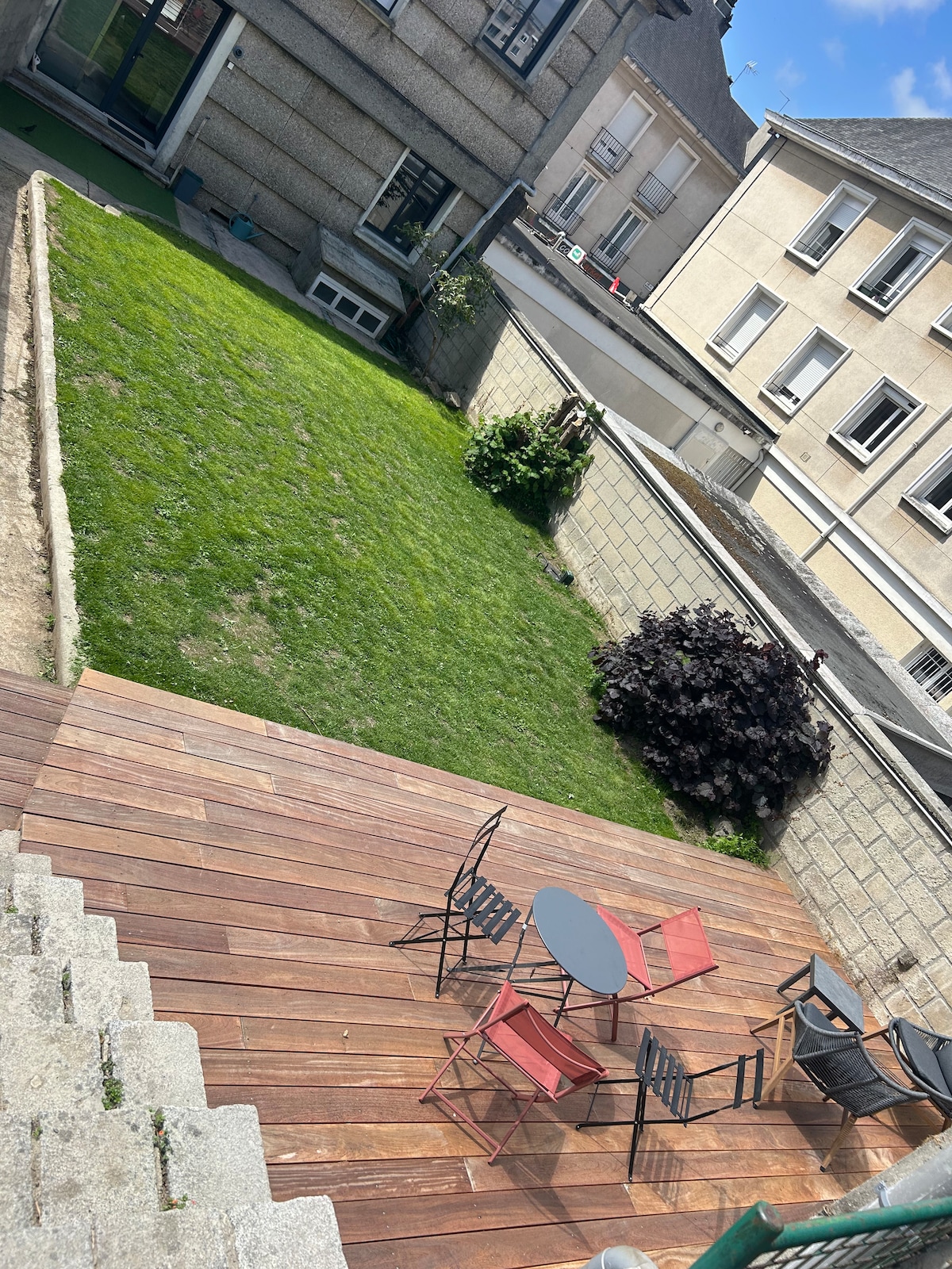 A wooden deck overlooks a well-maintained garden space. Four outdoor chairs surround a small circular table, with a grassy area extending beyond. The garden is enclosed by a stone wall, and various plants are visible within the greenery.