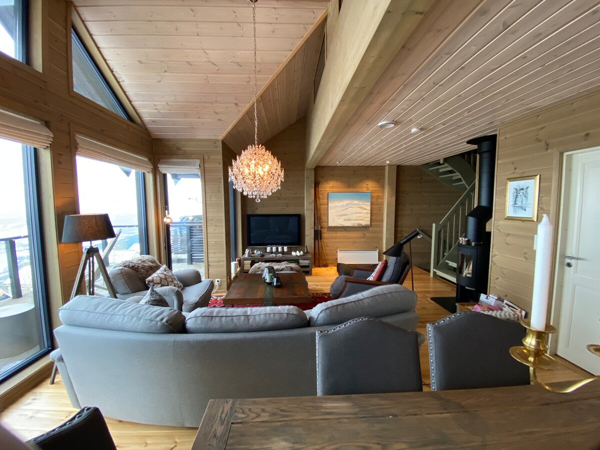 A spacious living area features large windows, allowing for ample natural light. A modern chandelier hangs above a comfortable seating arrangement, while a fireplace adds to the cozy ambiance. A staircase and decorative art can also be seen, contributing to the inviting atmosphere.