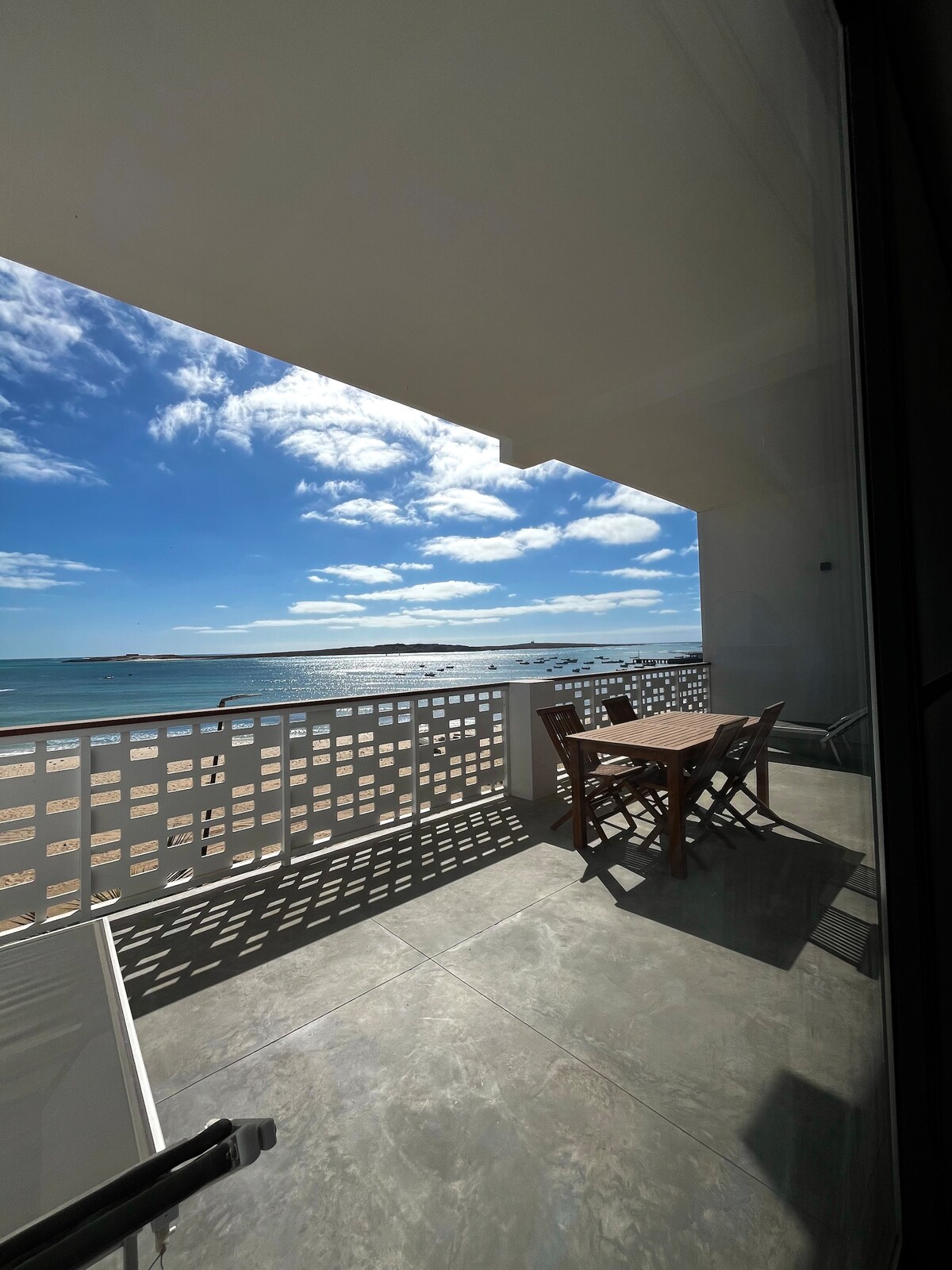 A spacious veranda is visible, featuring a wooden table and chairs overlooking the bay of Sal Rei. The open sky showcases scattered clouds, while sunlight casts gentle reflections on the water's surface.