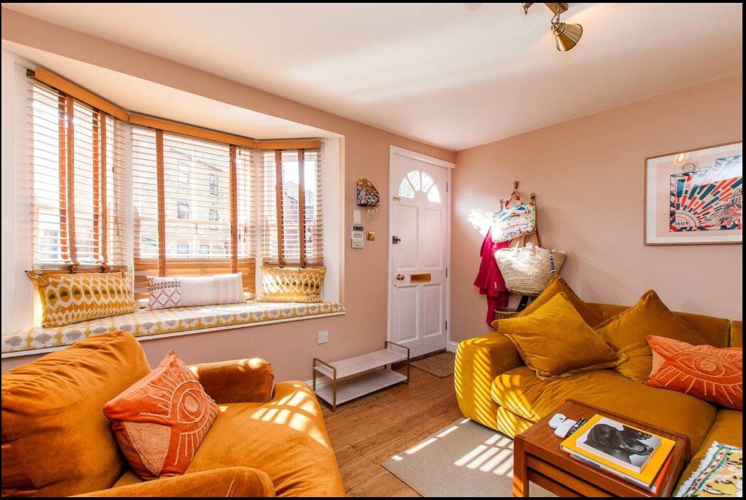 A welcoming living space features orange-hued seating and a bay window adorned with cushions. Natural light fills the room, highlighting the warm tones of the wooden floor. A door leads outside, and colorful artworks add character to the walls.
