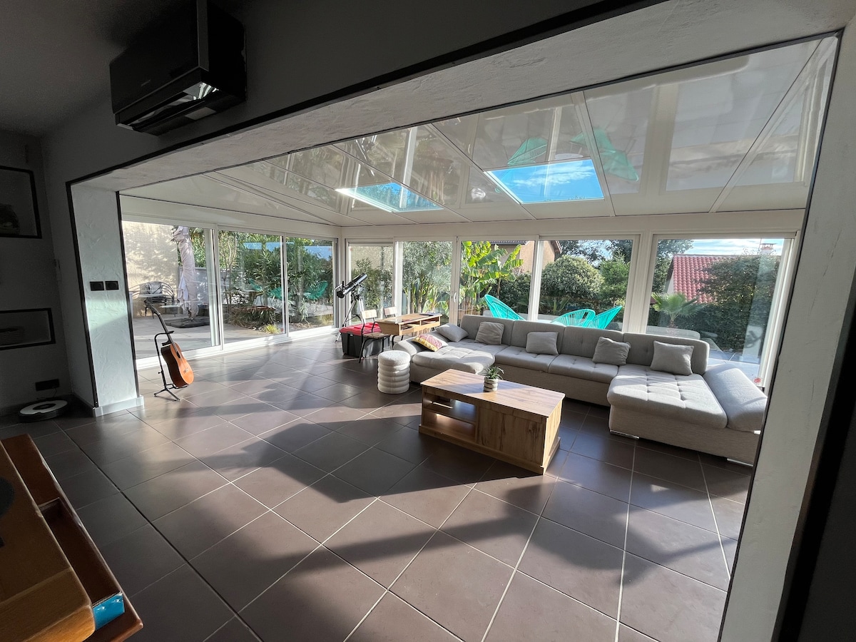 A spacious living area features large glass panels that provide ample natural light and outdoor views. A light-colored sectional sofa is positioned around a wooden coffee table. The area showcases a modern design with a mix of indoor greenery and elegant furnishings.