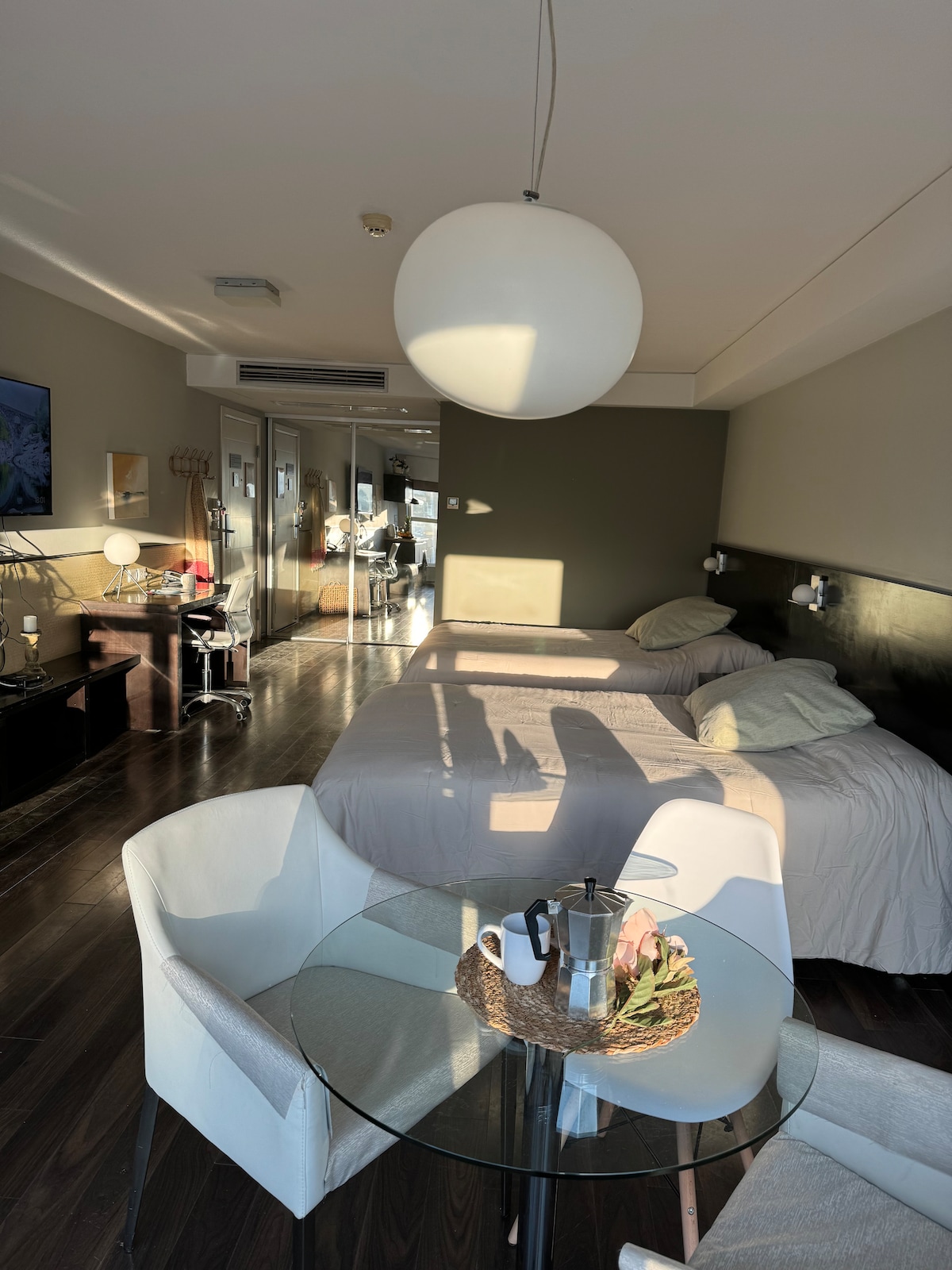 A spacious room is illuminated by natural light, featuring two beds topped with neutral-colored bedding. A round white pendant light hangs from the ceiling. A small glass dining table with a decorative tray sits beside a white armchair, creating a comfortable seating area.