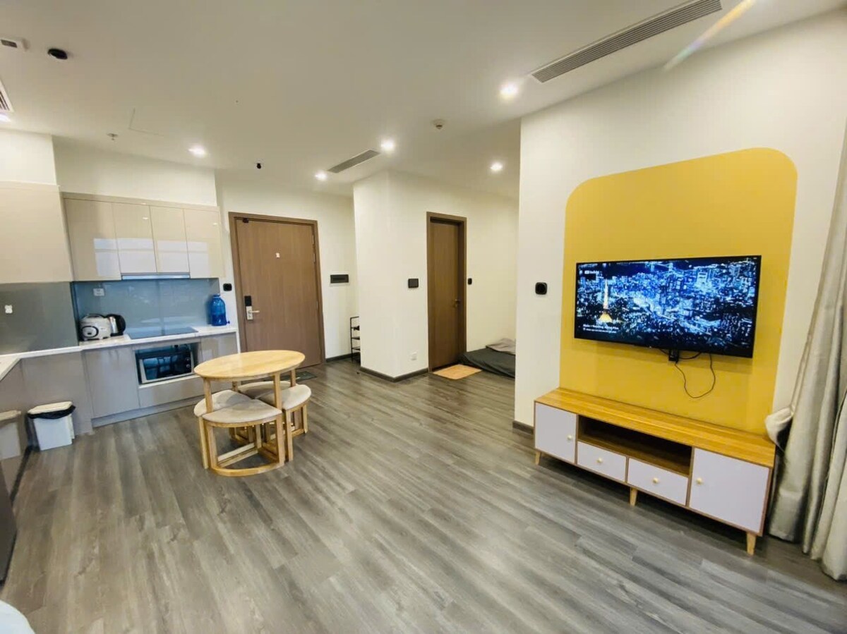 A modern living space features an open layout with a circular dining table accompanied by chairs. A television is mounted on the wall opposite a light-colored cabinet. The kitchen area showcases sleek cabinetry and appliances, while warm lighting enhances the interior, complemented by light wood flooring.