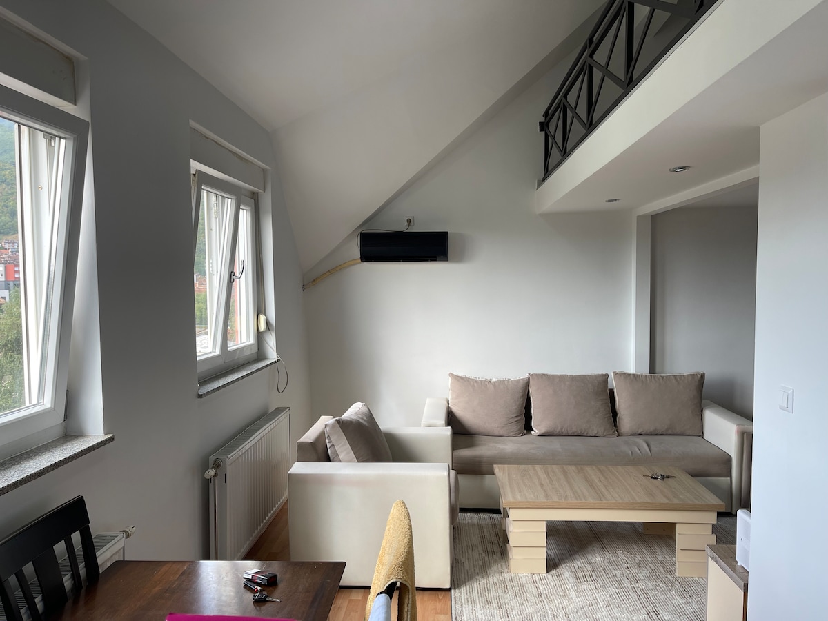 A bright living area features a comfortable light gray sofa, a wooden coffee table, and a neutral-tone area rug. Windows with soft curtains allow natural light to fill the space, while the high ceiling adds a sense of openness.