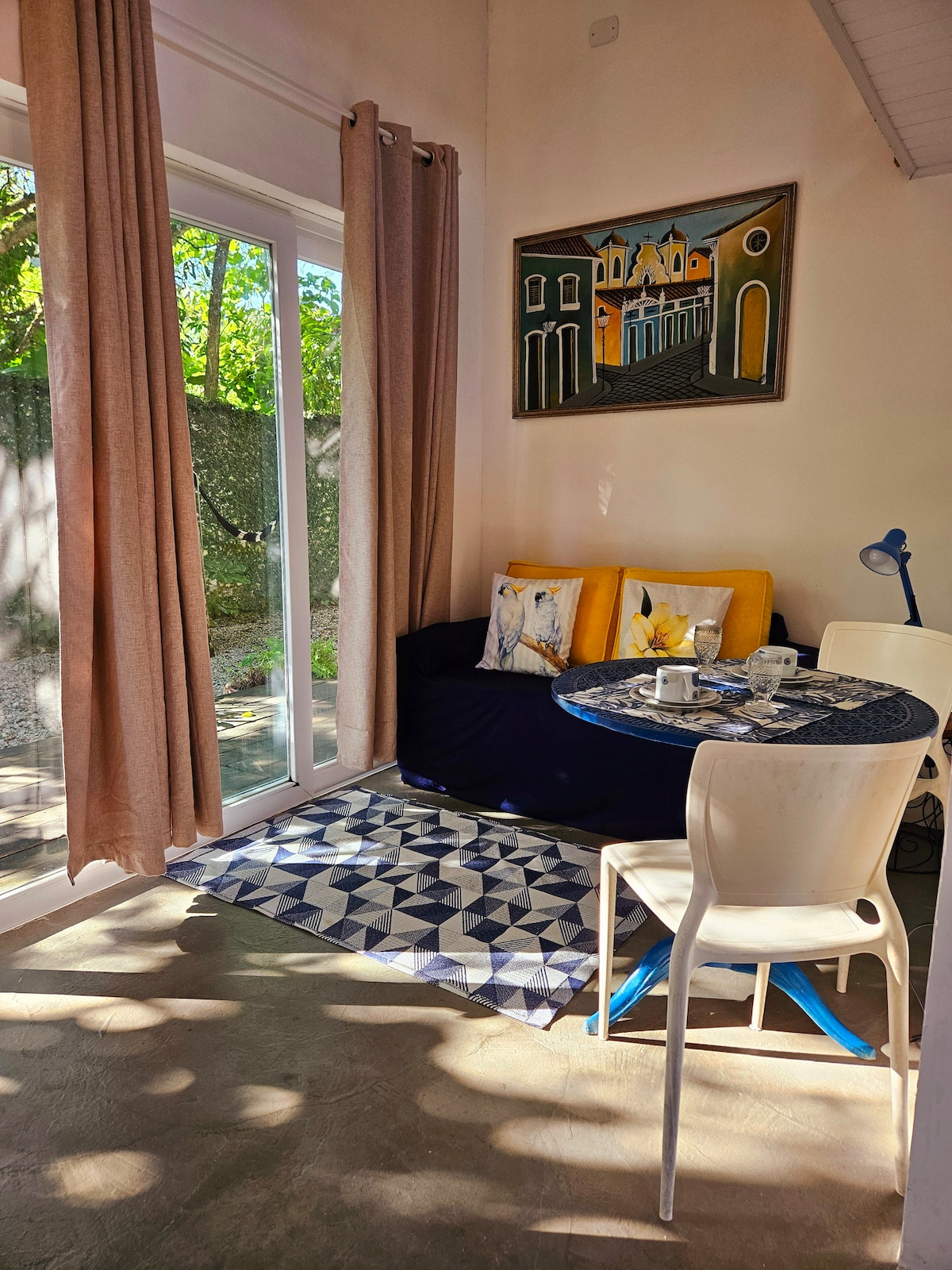 A cozy seating area is provided, featuring a dark sofa adorned with decorative pillows. A small dining table with white chairs is set, accompanied by a blue lamp. Natural light enters through large glass doors, illuminating the patterned rug and allowing views of the outside greenery.