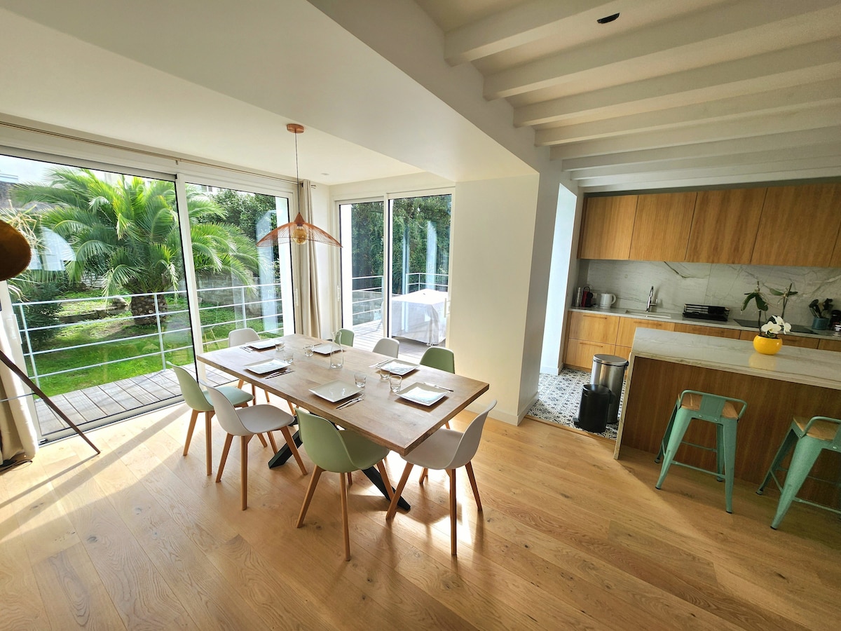 A spacious dining area features a wooden table set for meals, surrounded by light-colored chairs. Large windows provide a view of the lush garden, enhancing the natural light that fills the room. The open-plan kitchen is equipped with modern appliances, offering a seamless transition between spaces.