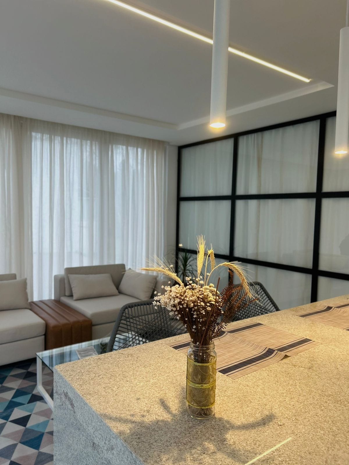 A modern open-concept living area features a light-colored sofa and wooden coffee table. Large windows allow natural light to fill the space, complemented by sheer white curtains. A decorative vase with dried flowers sits on the counter, enhancing the minimalist aesthetic.