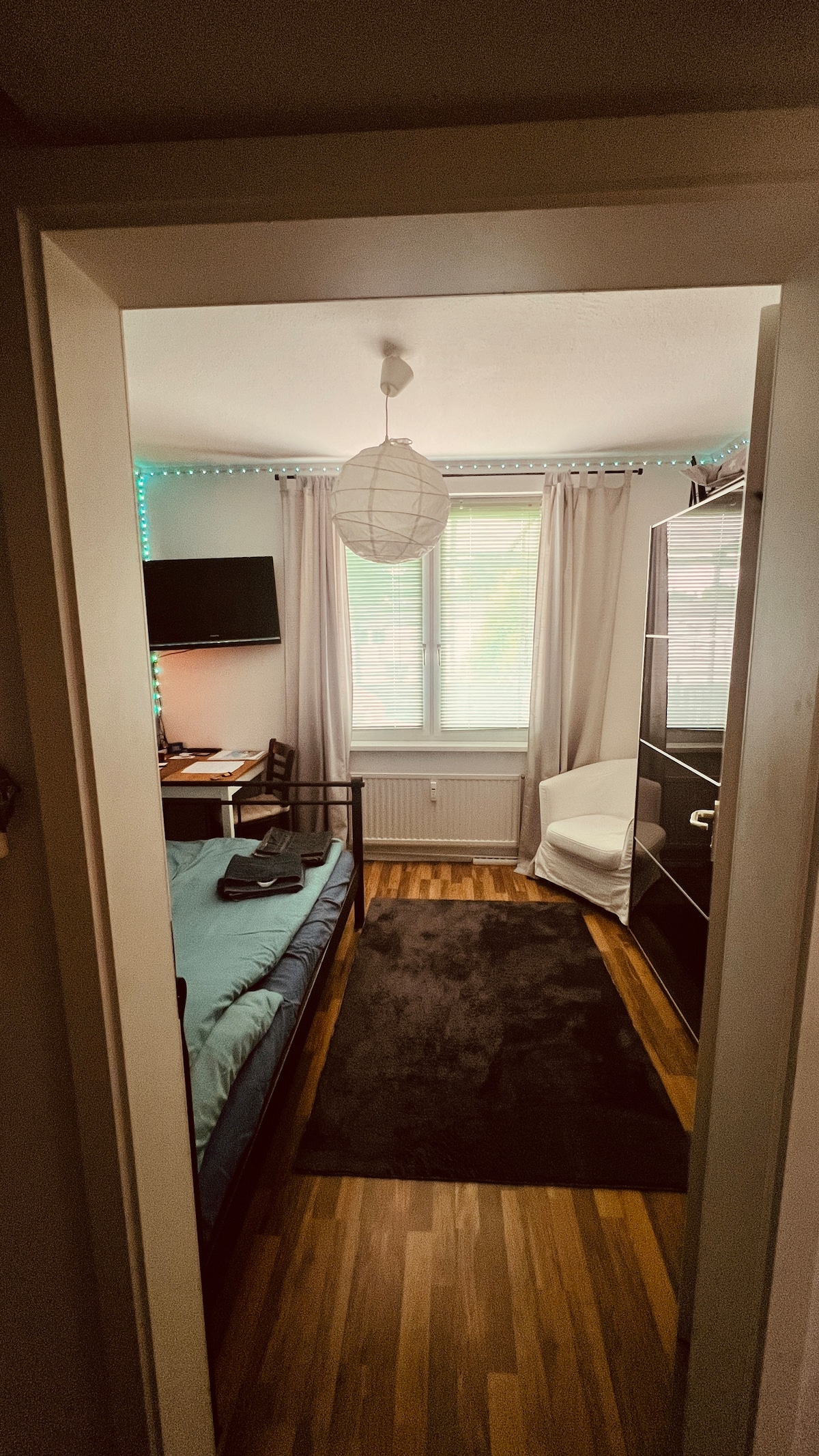 A bright room is displayed, featuring a spacious bed with a light blue cover. A small desk and chair are positioned near a window adorned with soft curtains. A cozy armchair is seen in one corner, and ambient lighting adds a gentle glow to the space.