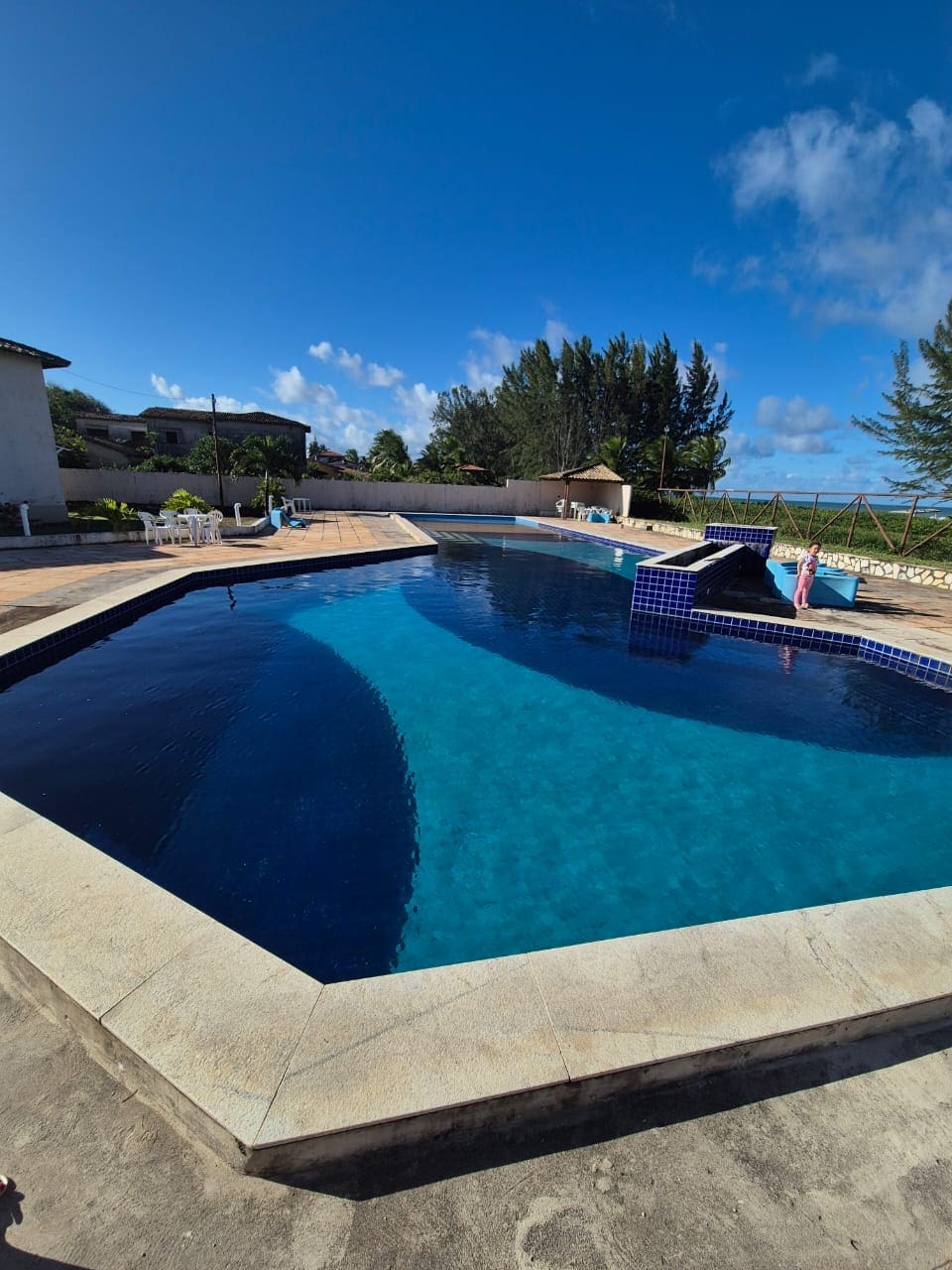 An inviting swimming pool with a vibrant blue surface is framed by a spacious pool deck. Lush greenery surrounds the area, and a water slide complements the outdoor space. The clear sky overhead enhances the atmosphere of relaxation.