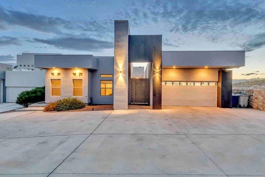A modern home's exterior is showcased, with clean lines and a striking facade. Warm lights accentuate the architectural features. The driveway leads up to a stylish garage door, while landscaped greenery adds a touch of nature to the contemporary design.