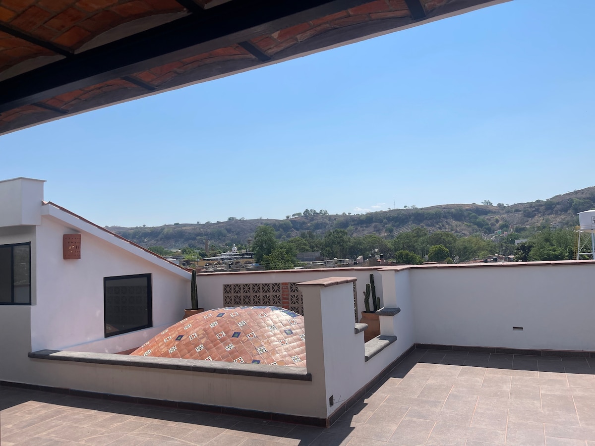 A spacious terrace is visible, featuring a tiled dome structure and potted cacti. A clear blue sky complements the natural scenery in the background, where rolling hills are seen. The terrace offers an open space ideal for relaxation and enjoying the surrounding landscape.