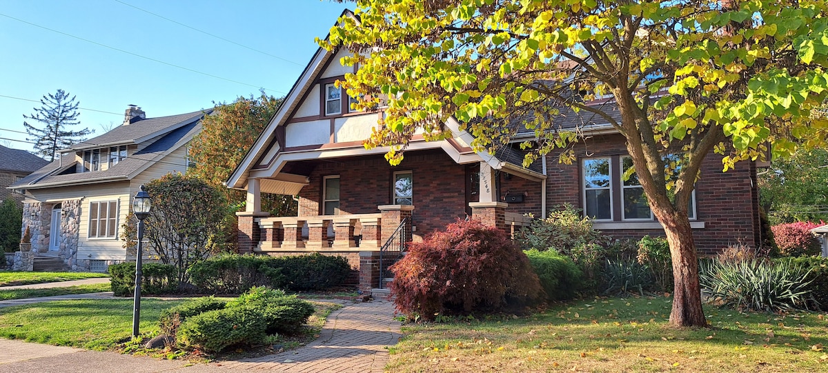 The exterior of a historic Craftsman home is showcased, featuring a welcoming porch with steps leading up to the entrance. The brick facade is complemented by lush greenery and well-maintained landscaping. A large tree provides shade, while colorful foliage adds to the tranquil ambiance.