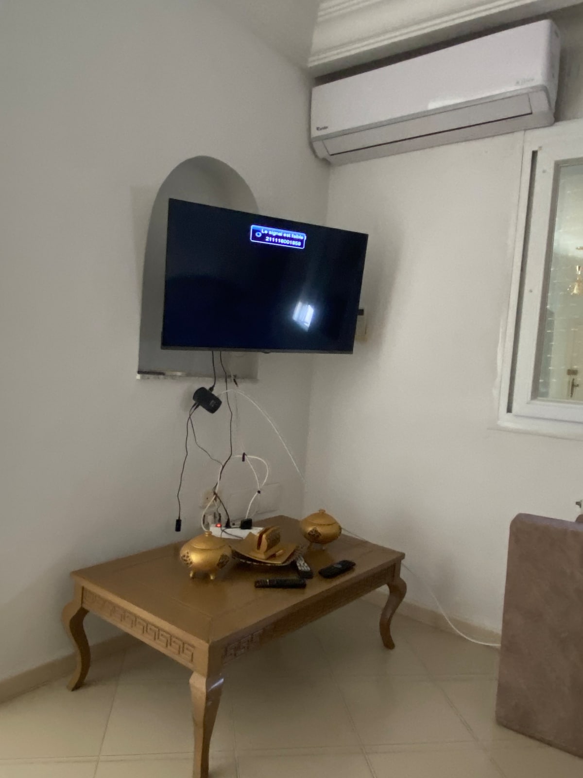 A wall-mounted television is positioned above a wooden table. The table displays two decorative items, with a remote control and other devices nearby. An air conditioning unit is mounted above, contributing to a comfortable environment.