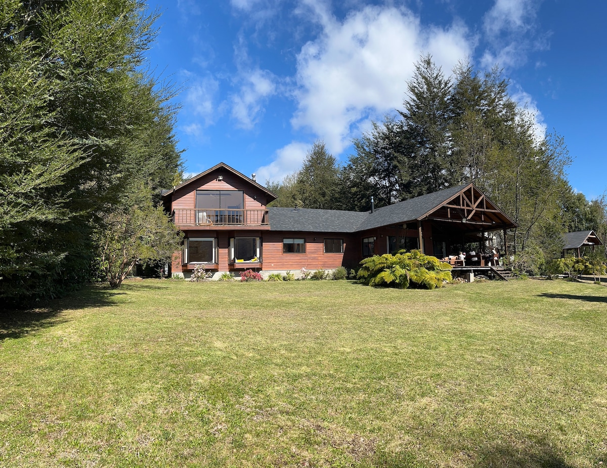 The exterior of a spacious house is surrounded by lush greenery and ornamental plants. A large deck extends from the house, offering a shaded area for relaxation. The sky is bright with scattered clouds, and a well-maintained lawn adds to the serene atmosphere.